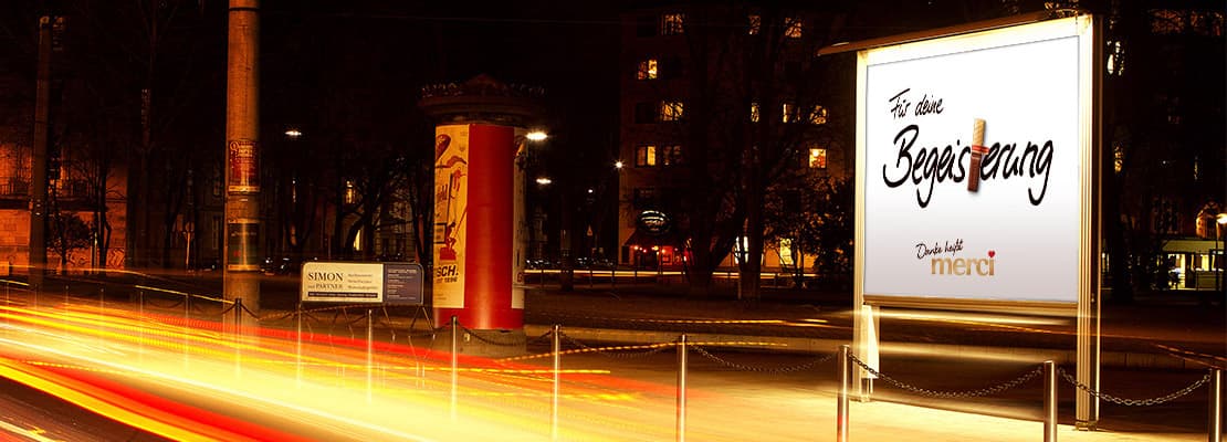 Beleuchtete Großfläche an einer Straße bei Nacht, im Hintergrund eine Litfaßsäule